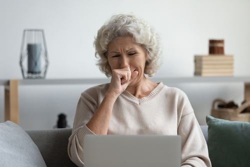 An older woman with gray hair sitting on a couch, looking at a laptop with a hand near her mouth, appearing sad and thoughtful.