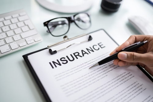 Close-up of a person reviewing an insurance policy document on a clipboard with a pen in hand, next to a keyboard and glasses on a desk.
