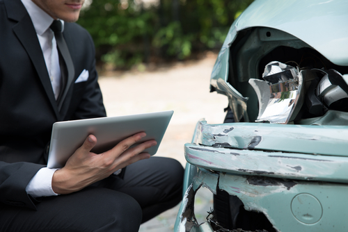 Man in a suit holding a tablet while inspecting damage to the front of a dented car.