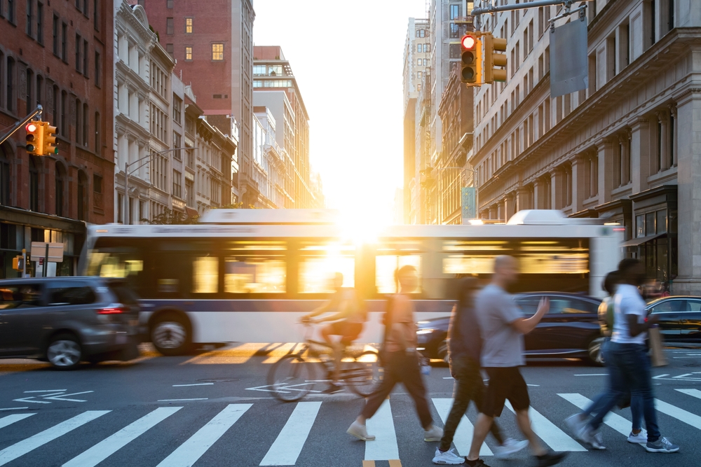 People, cars, bikes and buses traveling through a busy intersection on 5th Avenue and 23rd Street in New York City with sunlight shining in the background.