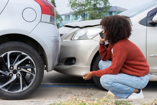 Young woman crouching to inspect damage after a rear-end collision, speaking on her phone beside two silver cars with dented bumpers.