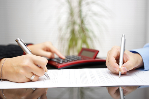Two people sign legal documents at a desk with a red calculator nearby, reflecting a business or legal agreement.