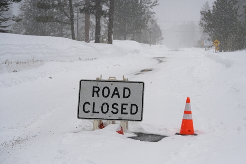 Road closed sign and traffic cone placed on a snow-covered road during heavy winter conditions.
