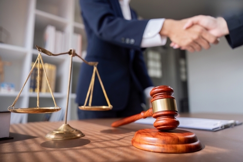 Close-up of a lawyer shaking hands with a client beside a wooden gavel and brass scales of justice, symbolizing a legal settlement.