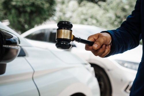 Close-up of a person in a blue suit holding a wooden gavel beside a white car, symbolizing legal action after a traffic accident.