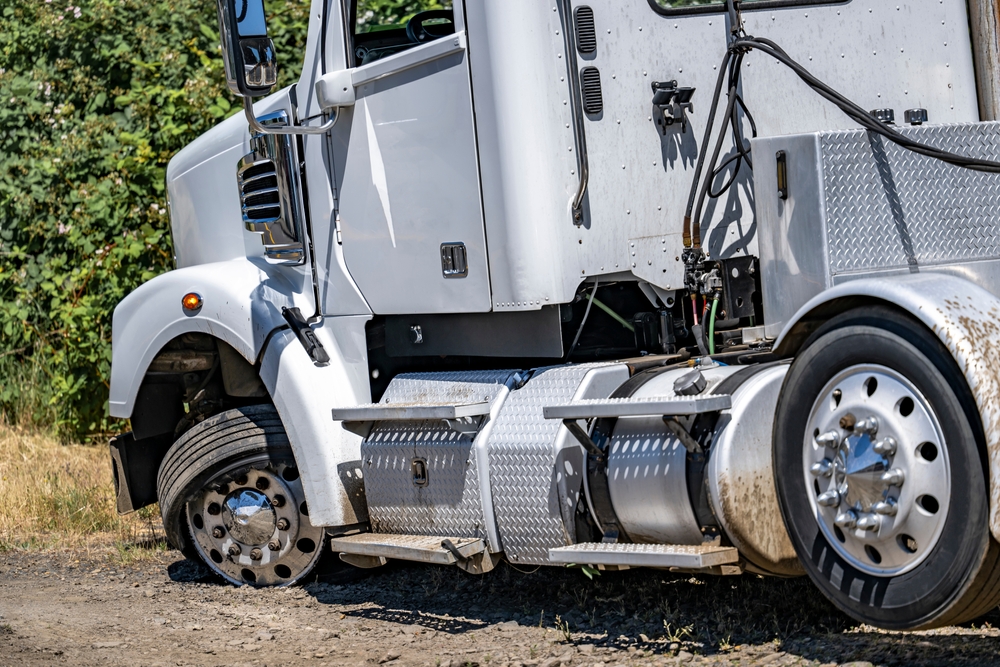 Industrial grade white big rig semi truck with a twisted wheel and a punctured tire standing out of service on the road shoulder waiting for mobile road assistant and tire repair technician