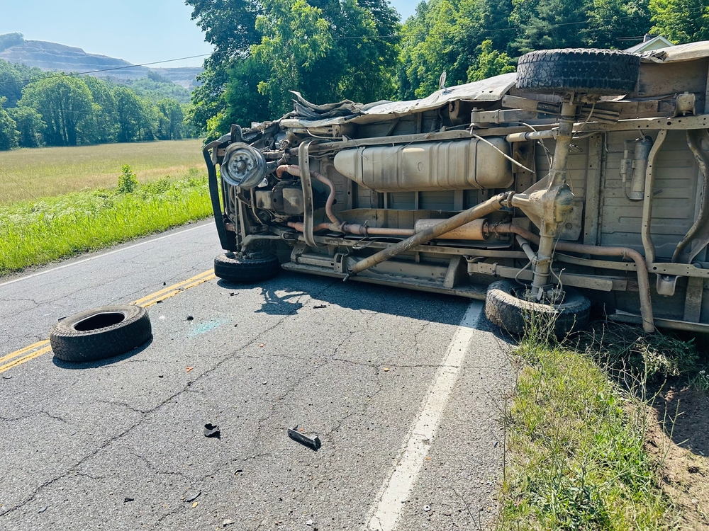 Photo of a motor vehicle accident scene where a cargo van was involved in a head on collision with a logging truck on a country road. The van was on its side and the truck was in a ditch.